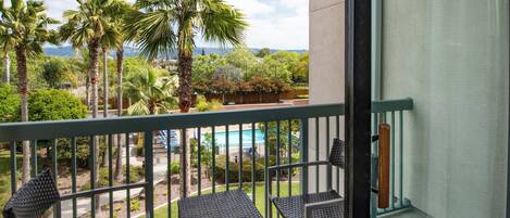 A balcony with two wicker chairs and a view of a swimming pool, palm trees, and a distant landscape.