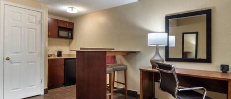 A hotel room with a kitchenette, a desk with a chair, a large mirror, and a door.