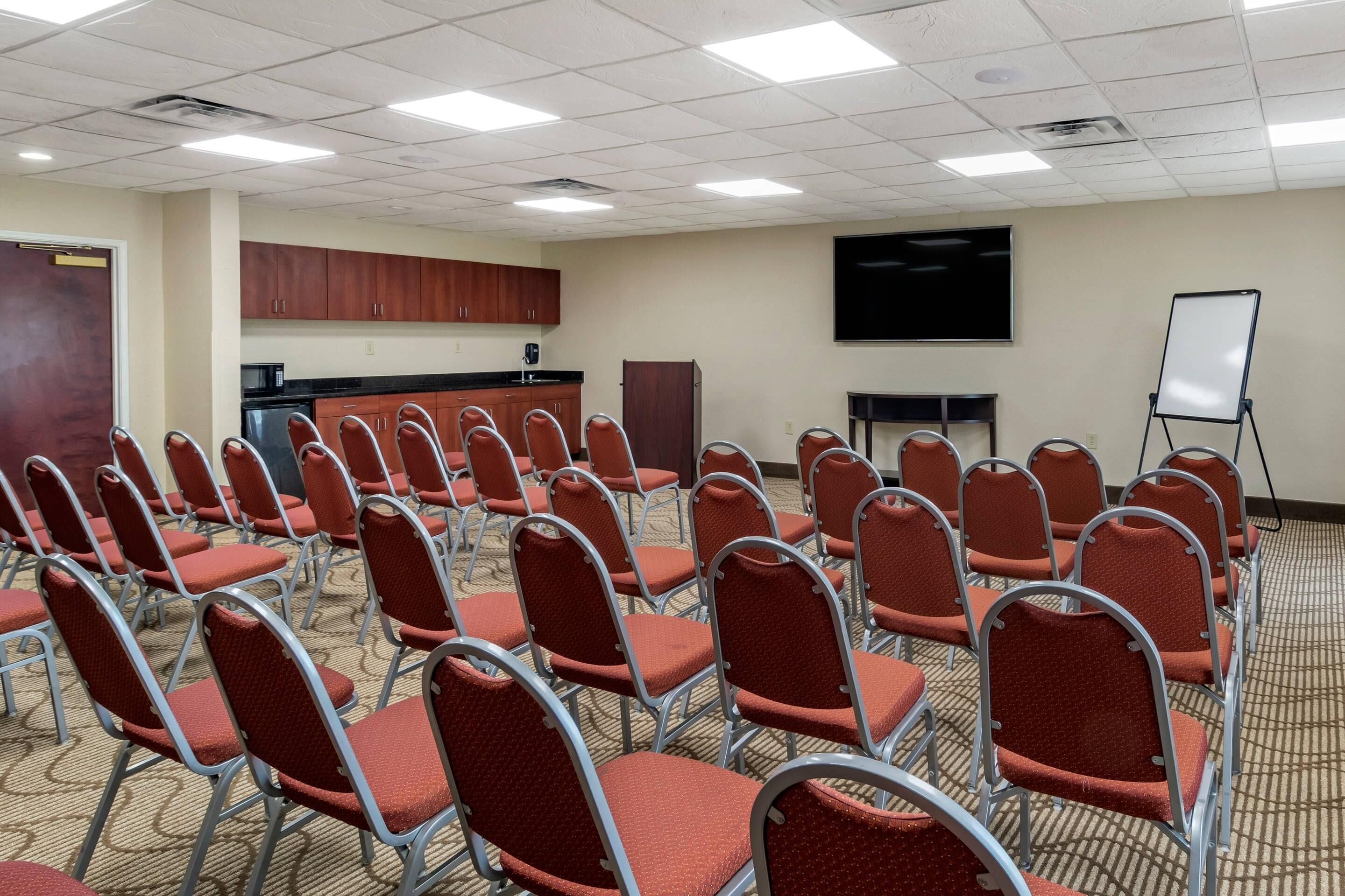 A conference room with rows of red chairs, a whiteboard, a projector screen, and a kitchenette area.