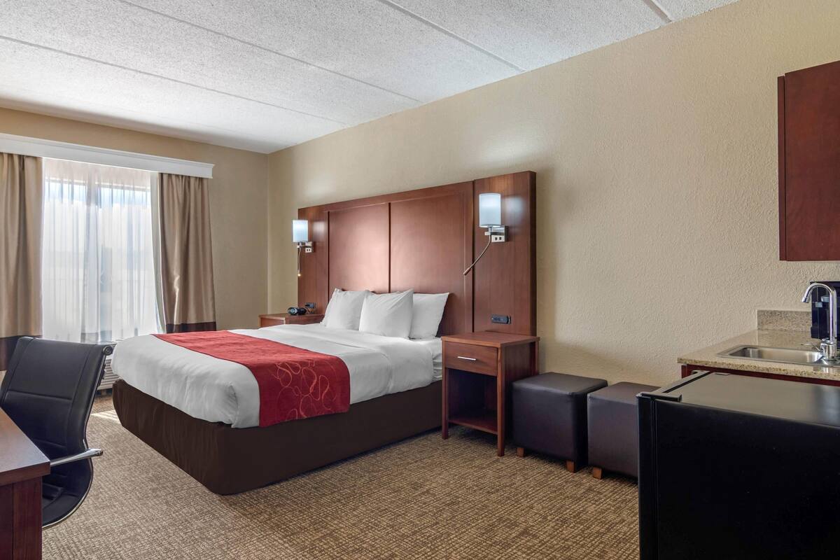 A hotel room with a bed, a desk, a chair, a small kitchenette, and a window with curtains.