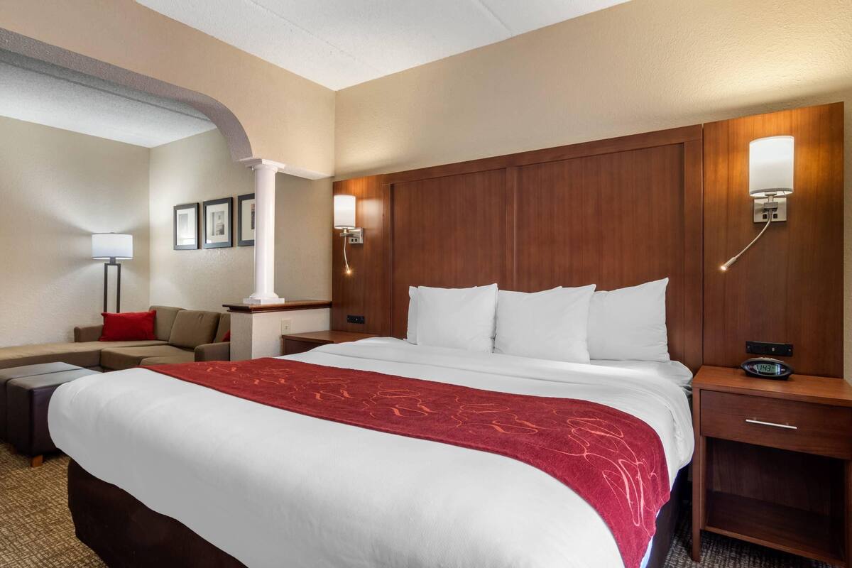 A hotel room with a large bed, a wooden headboard, a nightstand with a lamp, a desk, and a sofa.