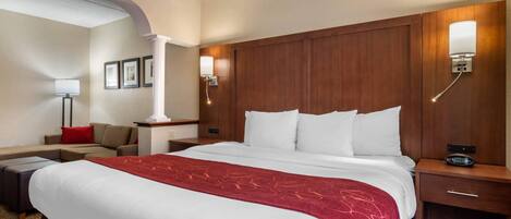 Habitación de hotel con una cama grande, un cabecero de madera, una mesita de noche con lámpara, un escritorio y un sofá.