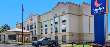 Un hotel con un letrero grande que dice 'Comfort Suites', una entrada cubierta y un asta con la bandera estadounidense.