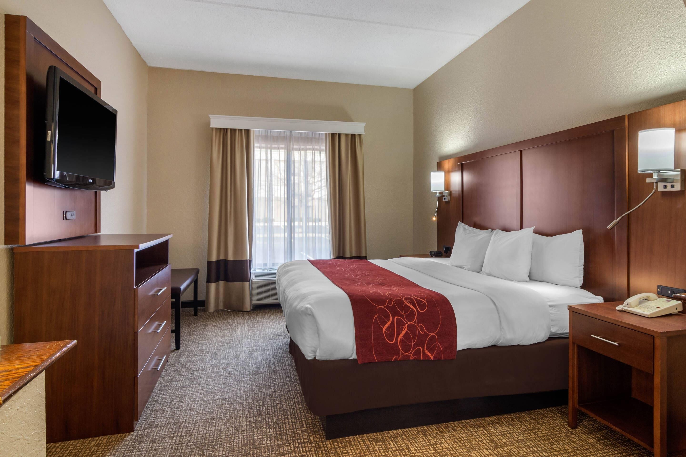 A hotel room with a bed, a desk, a television, a bedside table, and a window with curtains.