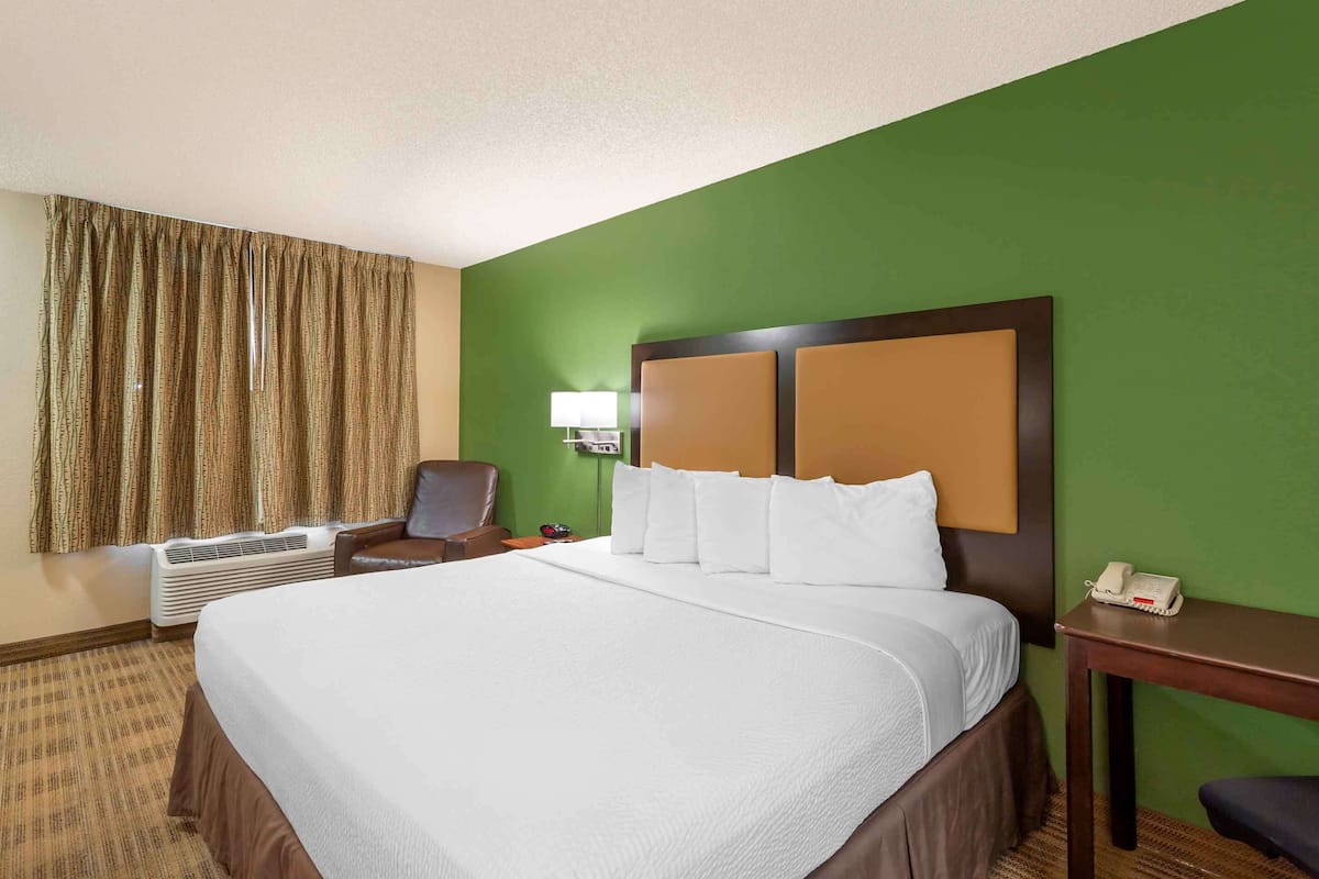 A hotel room with a large bed, a desk with a telephone, a chair, an air conditioning unit, and a window with curtains.