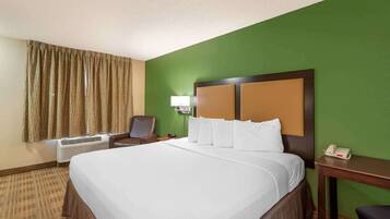 A hotel room with a large bed, a desk with a telephone, a chair, an air conditioning unit, and a window with curtains.