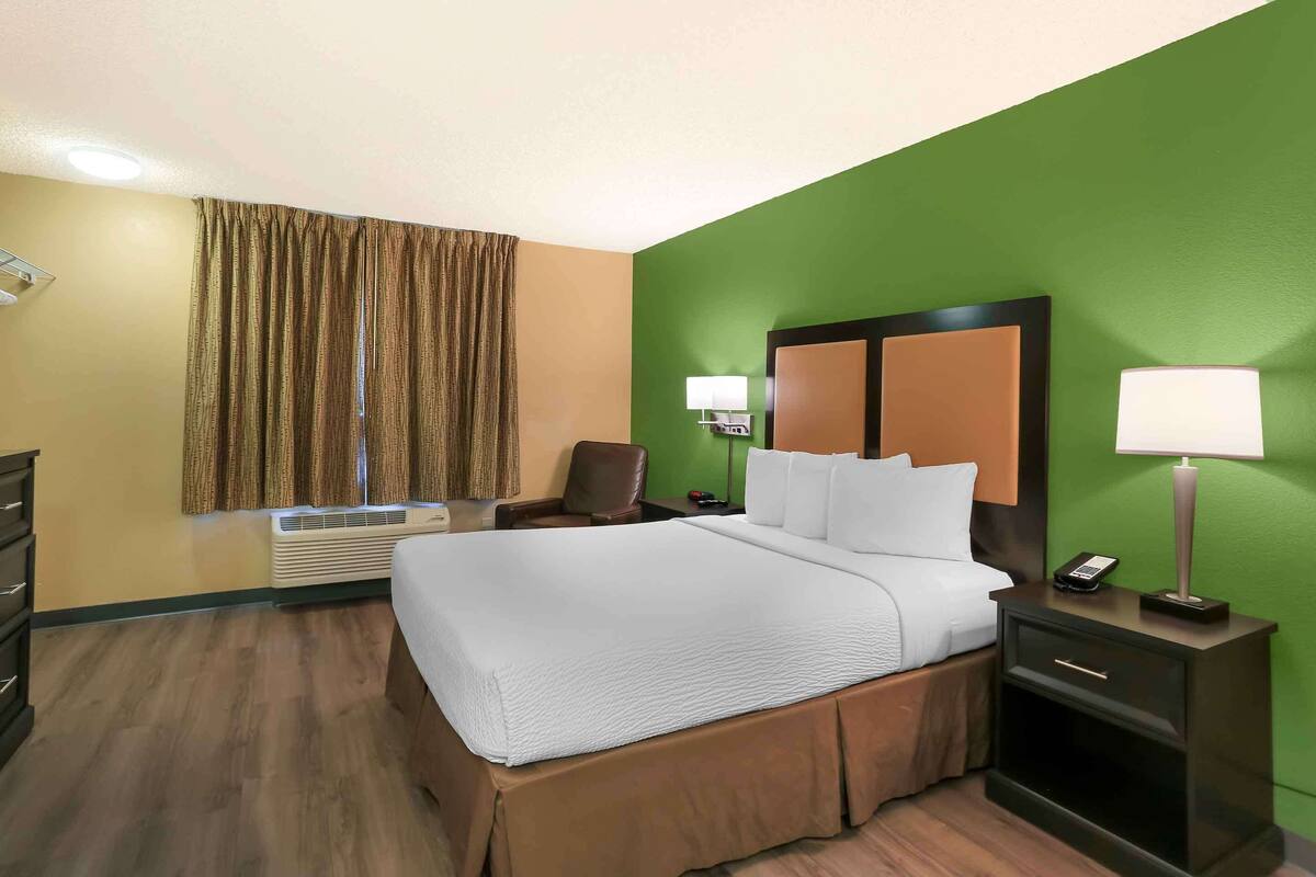 A hotel room with a large bed, a desk with a lamp, a chair, and a window with curtains.