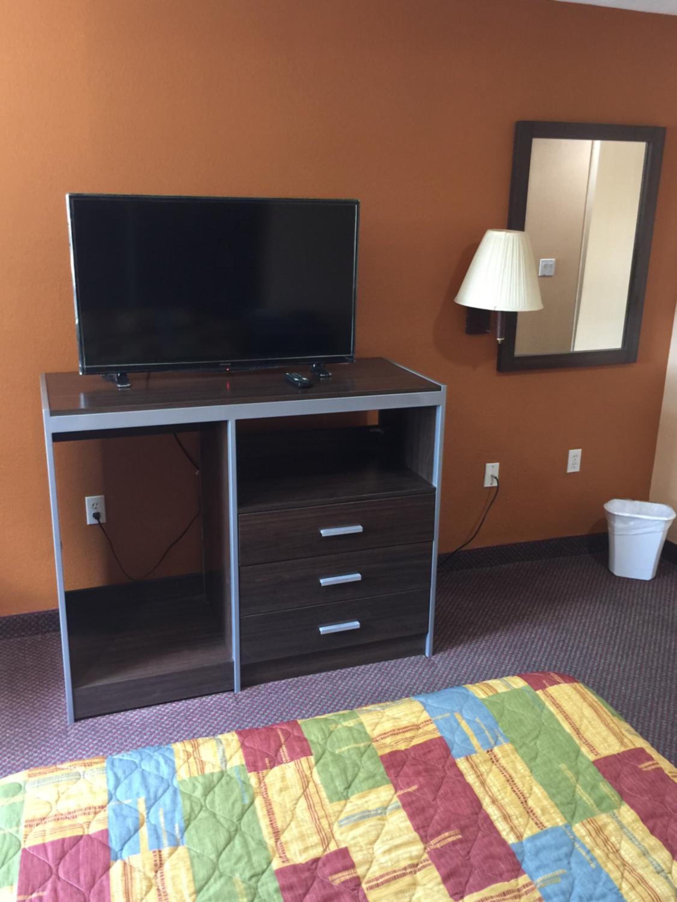 A hotel room with a desk, a television, a mirror, a lamp, a trash bin, and a colorful quilt on the bed.