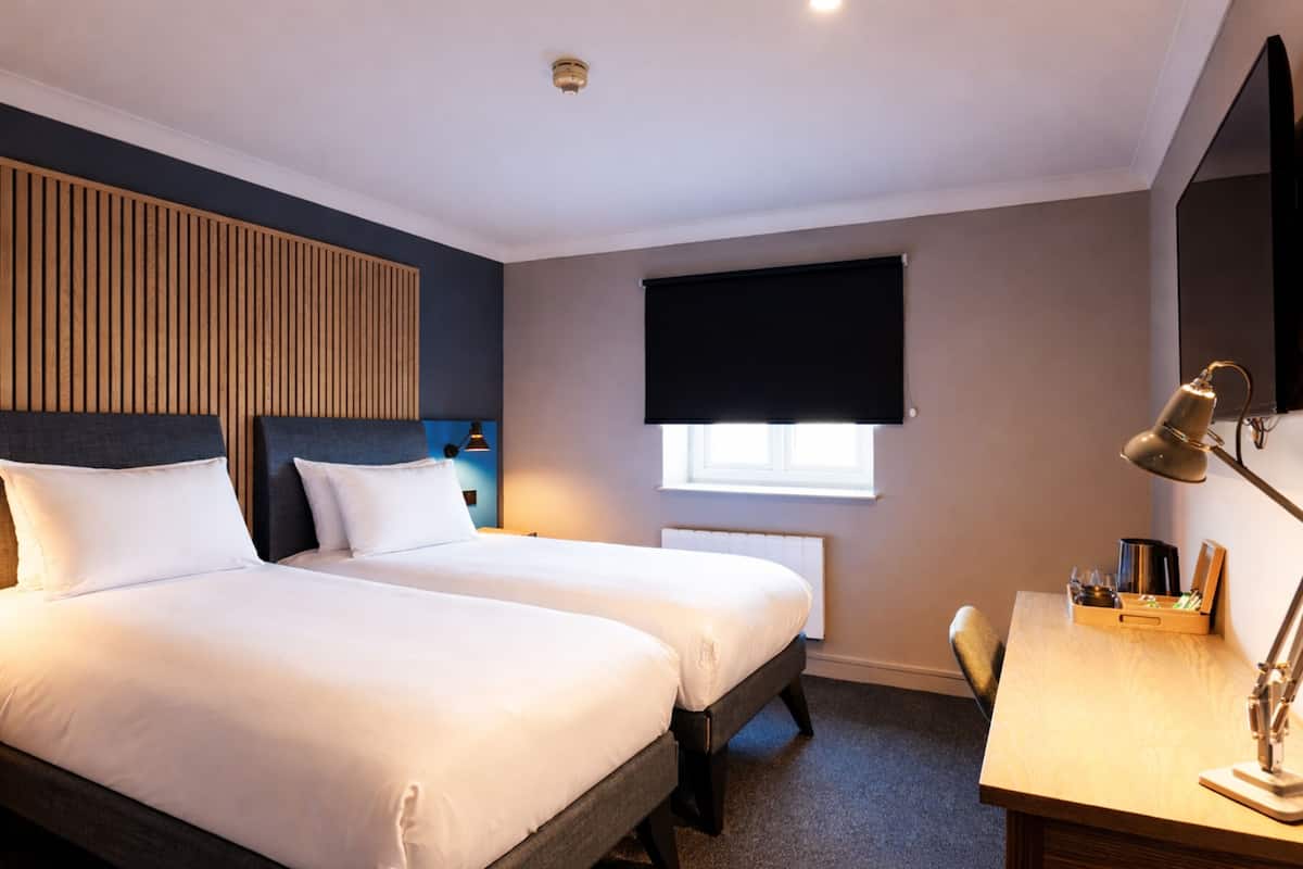 A hotel room with two beds, a desk, a chair, a window with a blind, and a radiator.