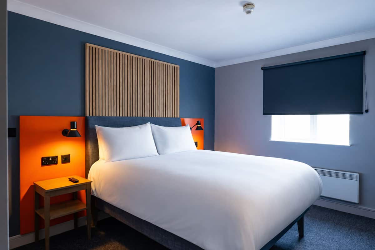 A hotel room with a bed, a nightstand, a wall-mounted lamp, a window with a blind, and a radiator.