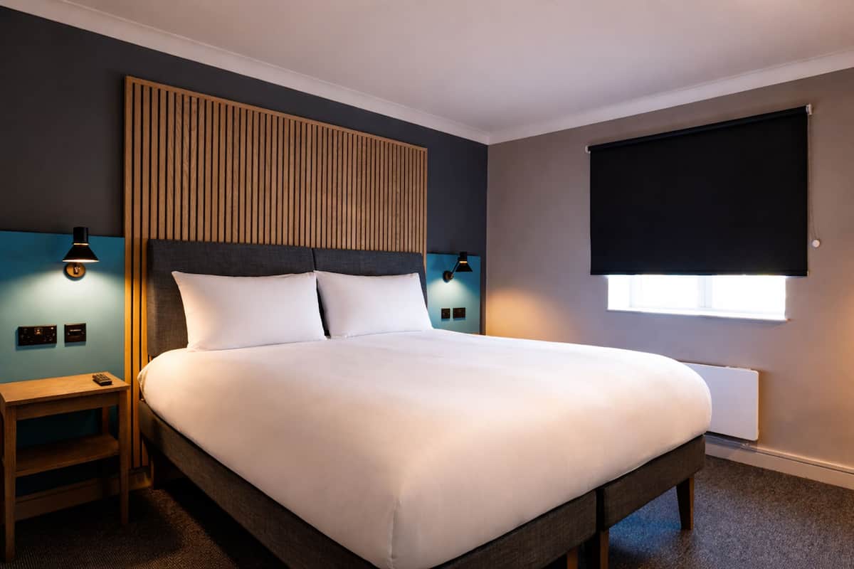 A hotel room with a large bed, wooden headboard, two bedside tables with lamps, a window with a blind, and a wall-mounted thermostat.