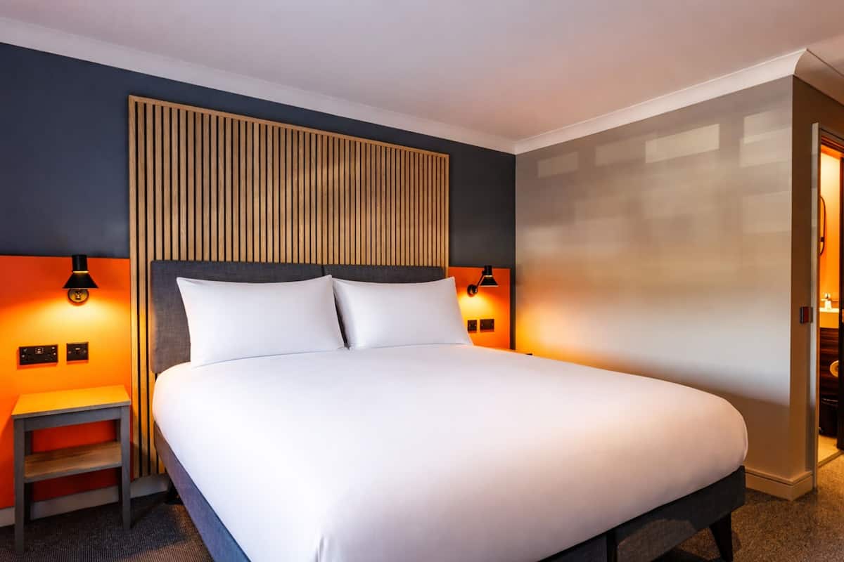A hotel room with a large bed, bedside lamps, a small table, and a wooden headboard.