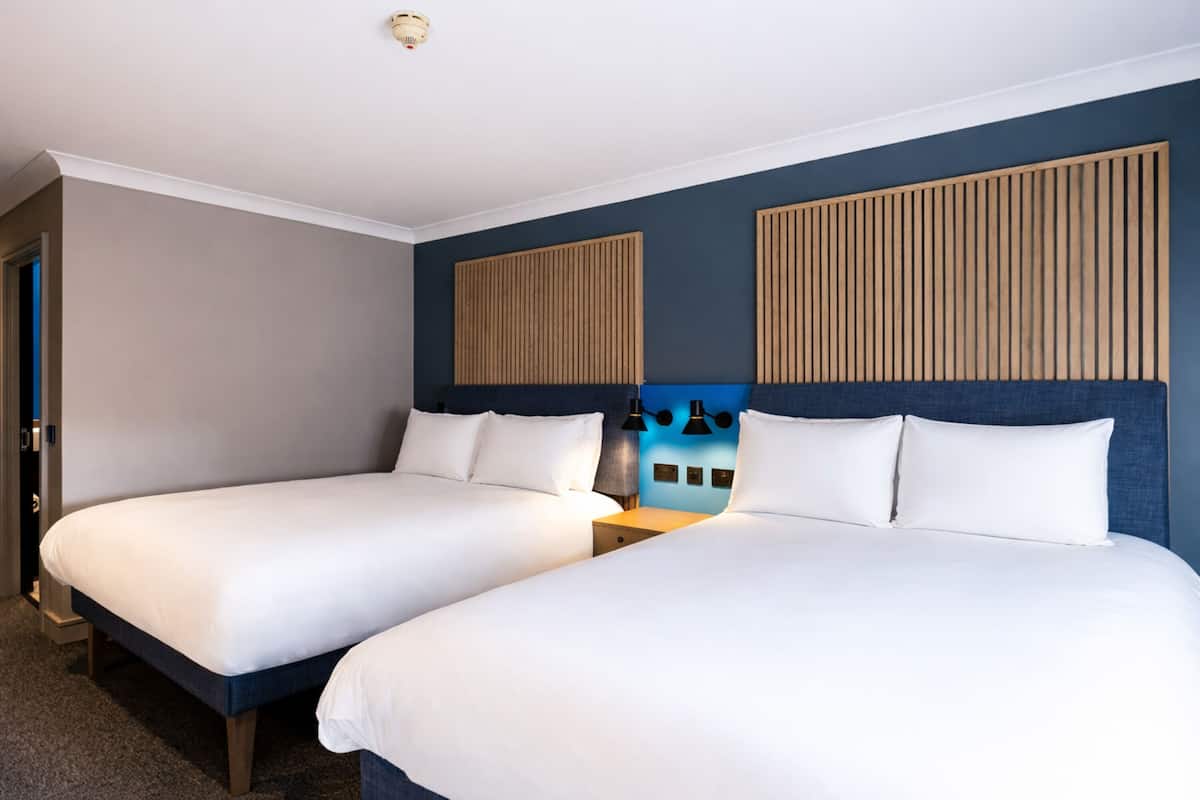 A hotel room with two beds, a wooden headboard, a nightstand, and a wall-mounted light fixture.