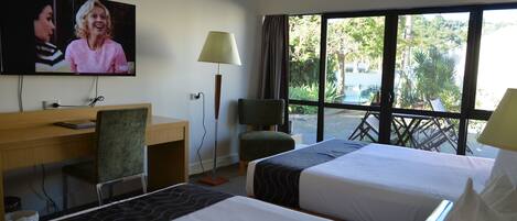 A hotel room with two beds, a desk, a TV, and a window with a view of greenery.
