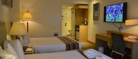 Quarto de hotel com duas camas, uma escrivaninha, televisão e casa de banho visível através de uma porta aberta.