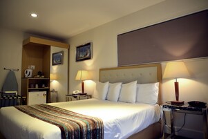 A hotel room with a large bed, two bedside lamps, a desk with a telephone, a small refrigerator, a mirror, and a picture on the wall.