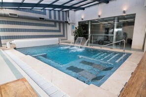 An indoor swimming pool with a waterfall feature, surrounded by a tiled floor and a glass wall. - Hotel Spa Paris (León)