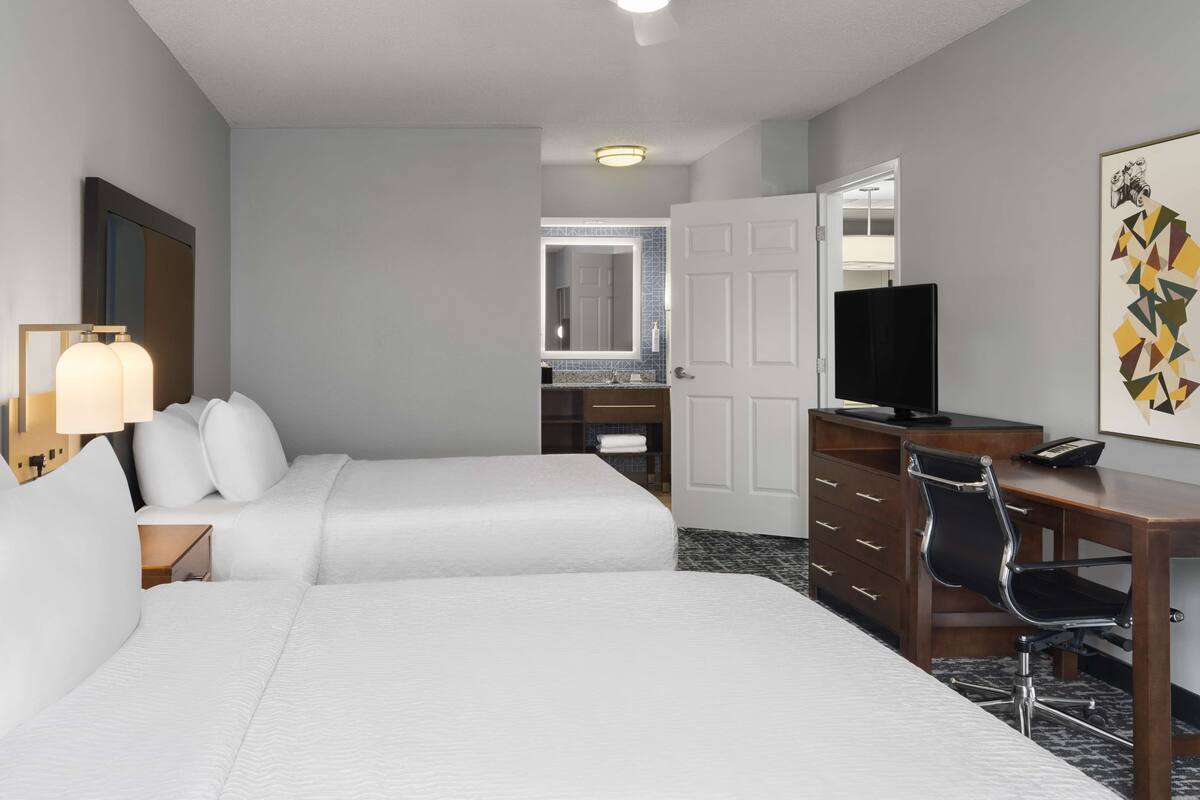 Une chambre d’hôtel avec deux lits, un bureau avec une chaise, une télévision et une salle de bain visible à travers une porte ouverte.