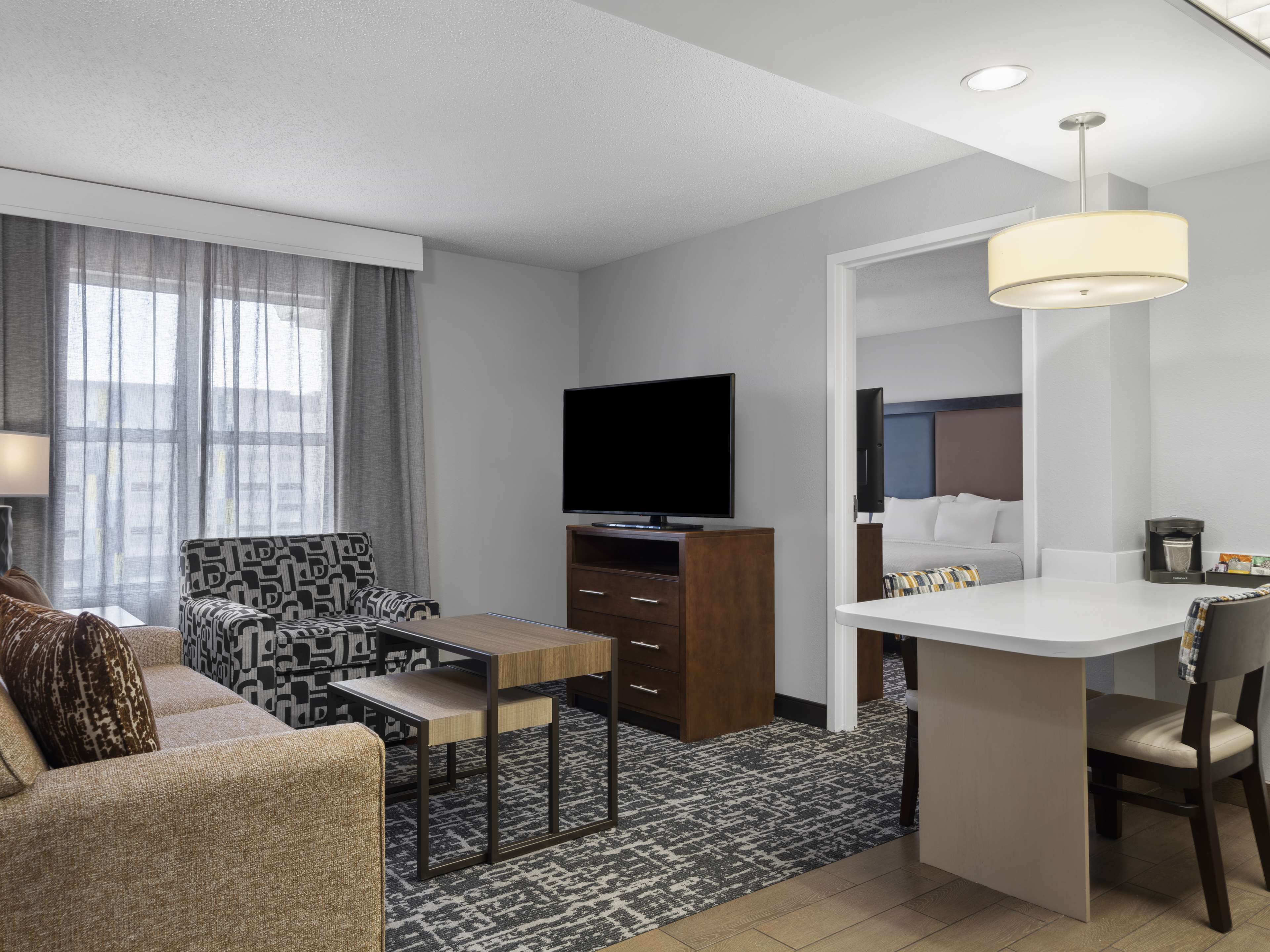 A modern hotel room with a sofa, a small table, a TV, a desk with a chair, and a large mirror.