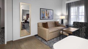A hotel room with a sofa, a small table, a mirror, a framed cityscape artwork, and a window with curtains.