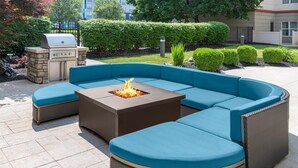 A sectional sofa with a square fire pit in the center, surrounded by a stone barbecue grill, set on a paved patio with a landscaped garden in the background.