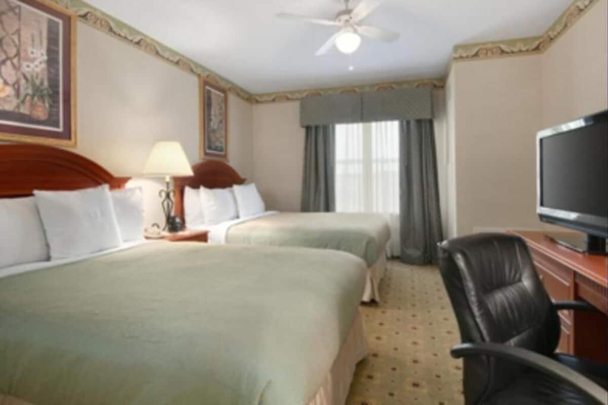 Une chambre d’hôtel avec deux lits, un bureau avec une télévision, une chaise, un ventilateur de plafond et une fenêtre avec des rideaux.