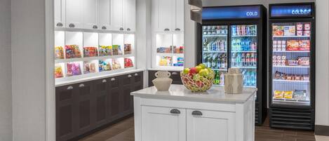 A clean, well-lit store with white cabinets and a central island. There are refrigerated sections labeled 'COLD' and 'COLDER' with various beverages and snacks.