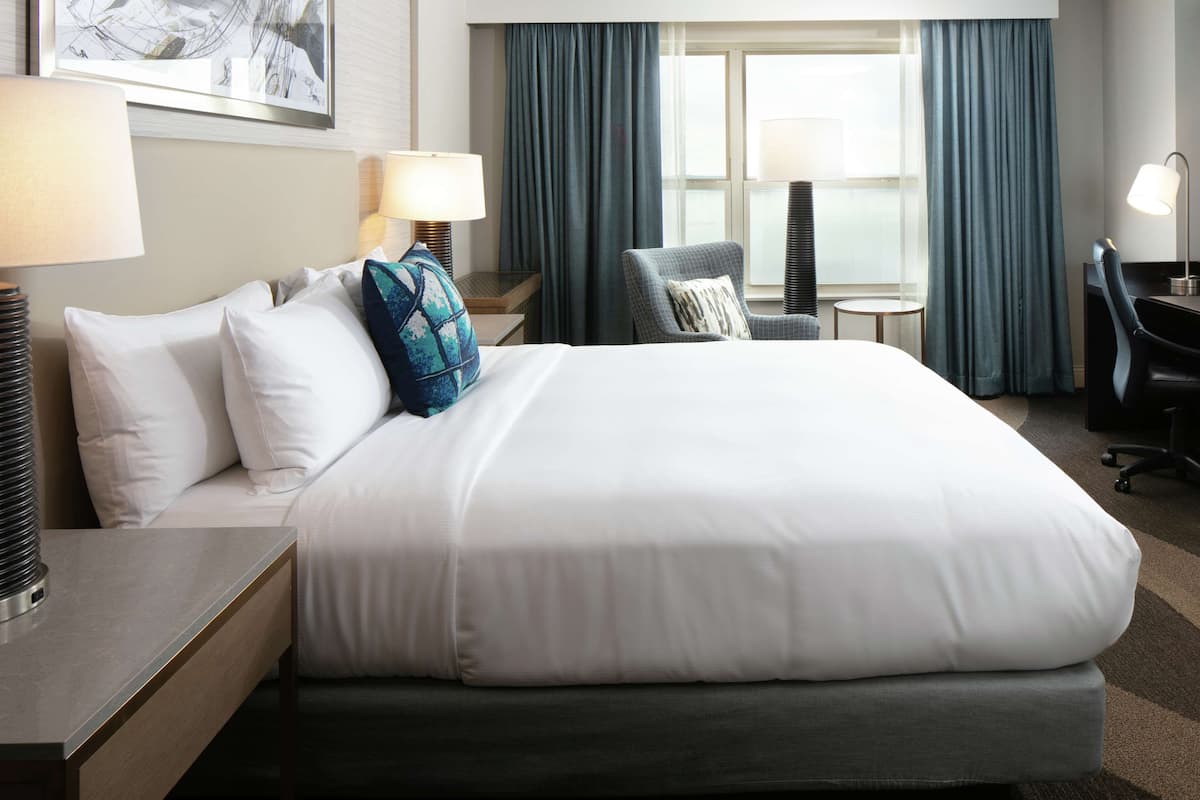 A hotel room with a large bed, a desk with a lamp, a chair, and a view of the outdoors through a window.