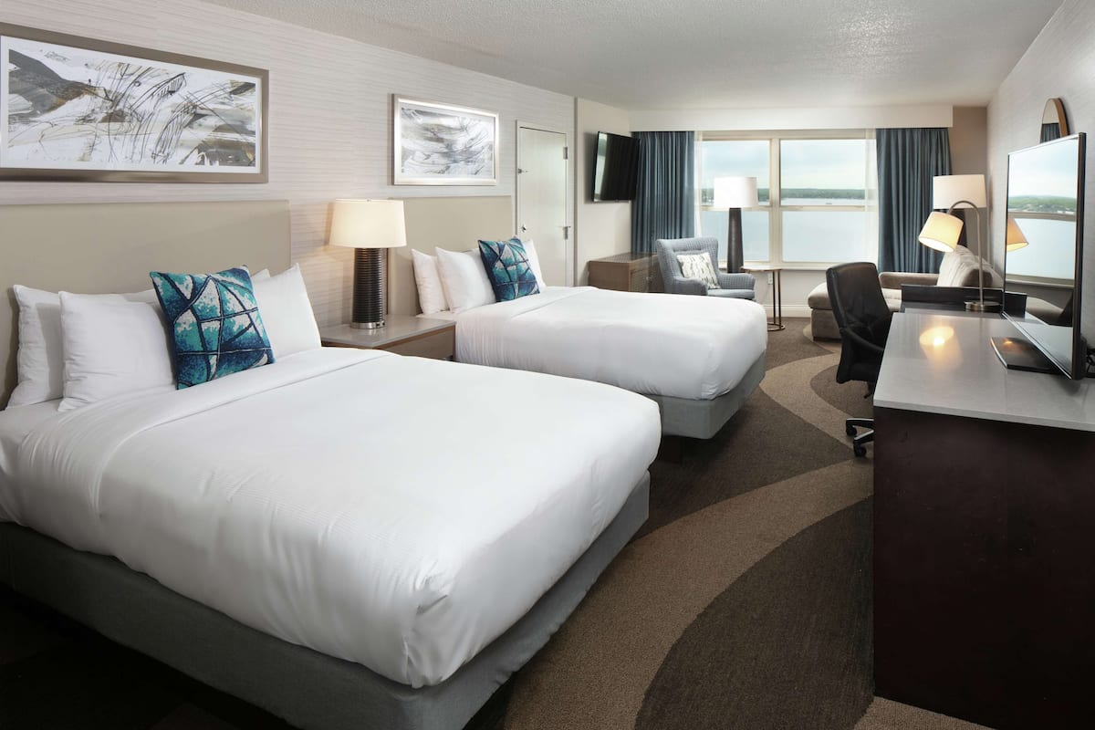 A hotel room with two beds, a desk, a chair, a TV, and a large window with a view.