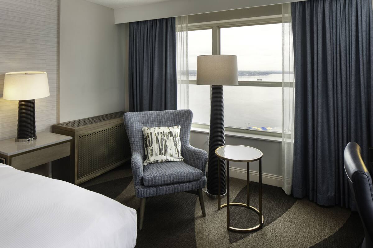 A hotel room with a bed, a blue armchair with a patterned pillow, a standing lamp, a small round table, and a view of the water through large windows.