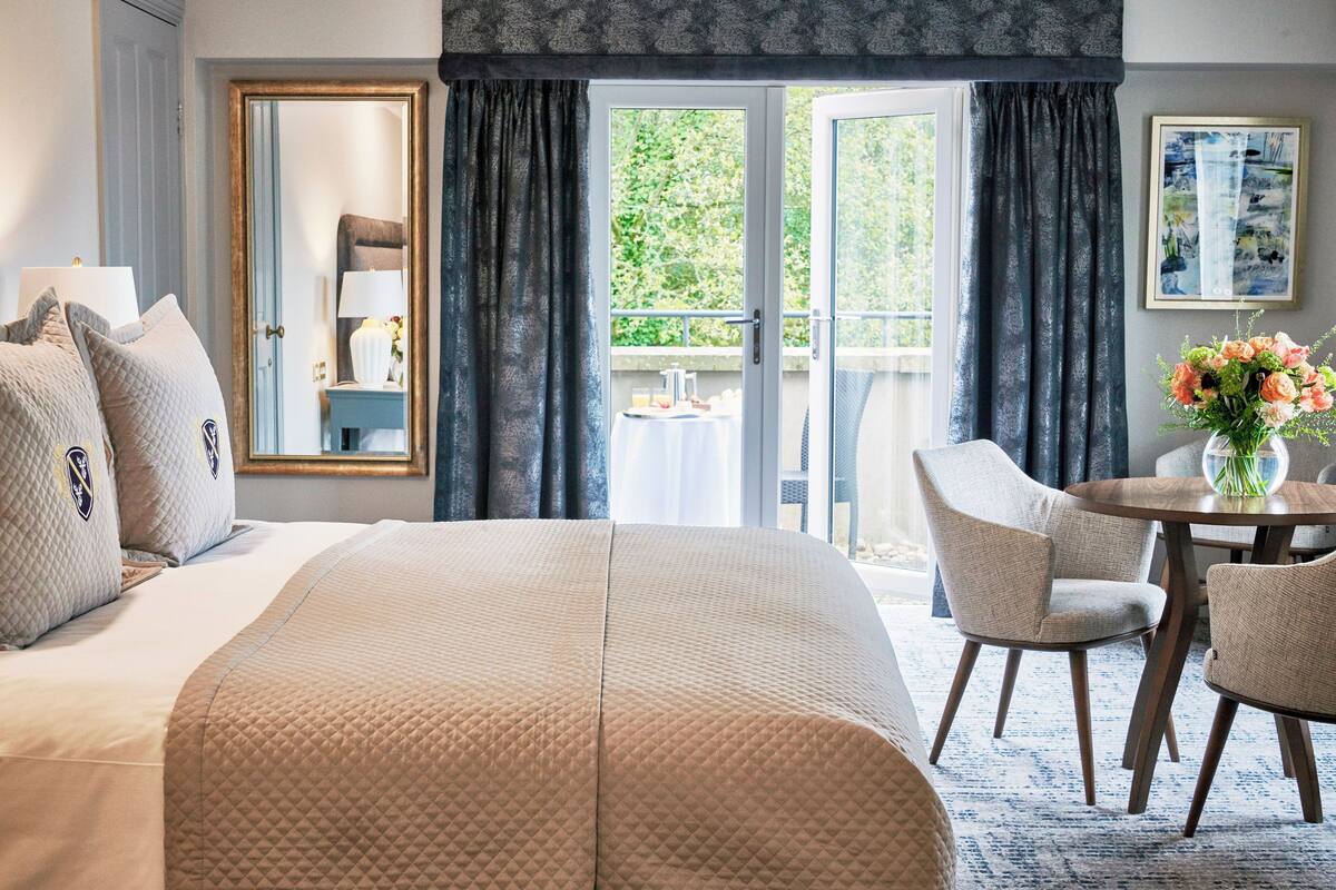 A hotel room with a large bed, a sitting area with a table and chairs, a balcony with a view of greenery, and a painting on the wall.