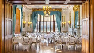 A formal dining room with multiple tables set for a meal, large windows, and a chandelier.