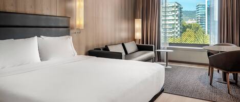 A modern hotel room with a large bed, a sofa, a small table, and a view of the cityscape through large windows.