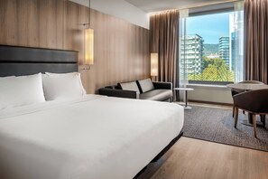 A modern hotel room with a large bed, a sofa, a small table, and a view of the cityscape through large windows.