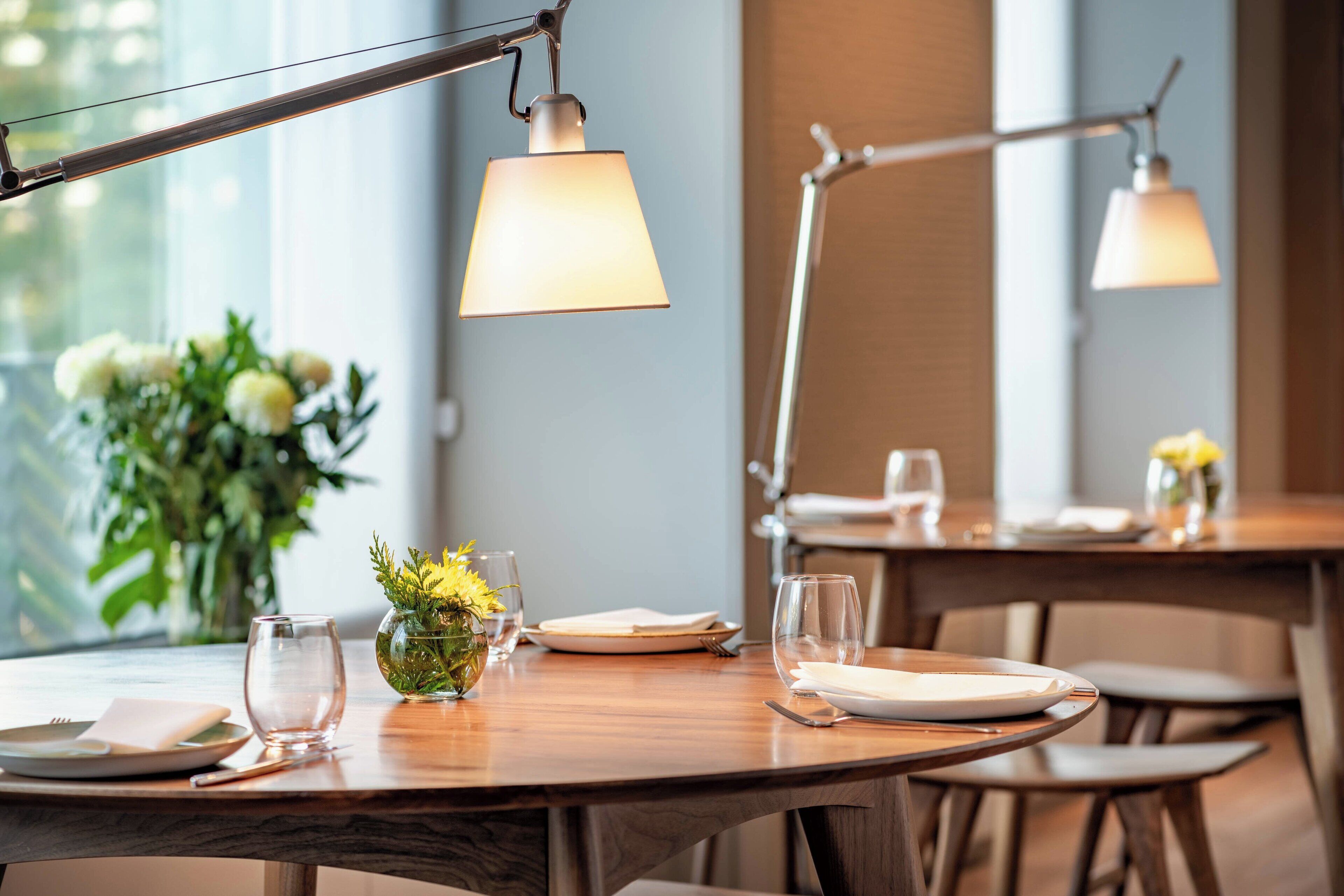 A dining table set with plates, glasses, and cutlery, with a vase of flowers and a lamp overhead.