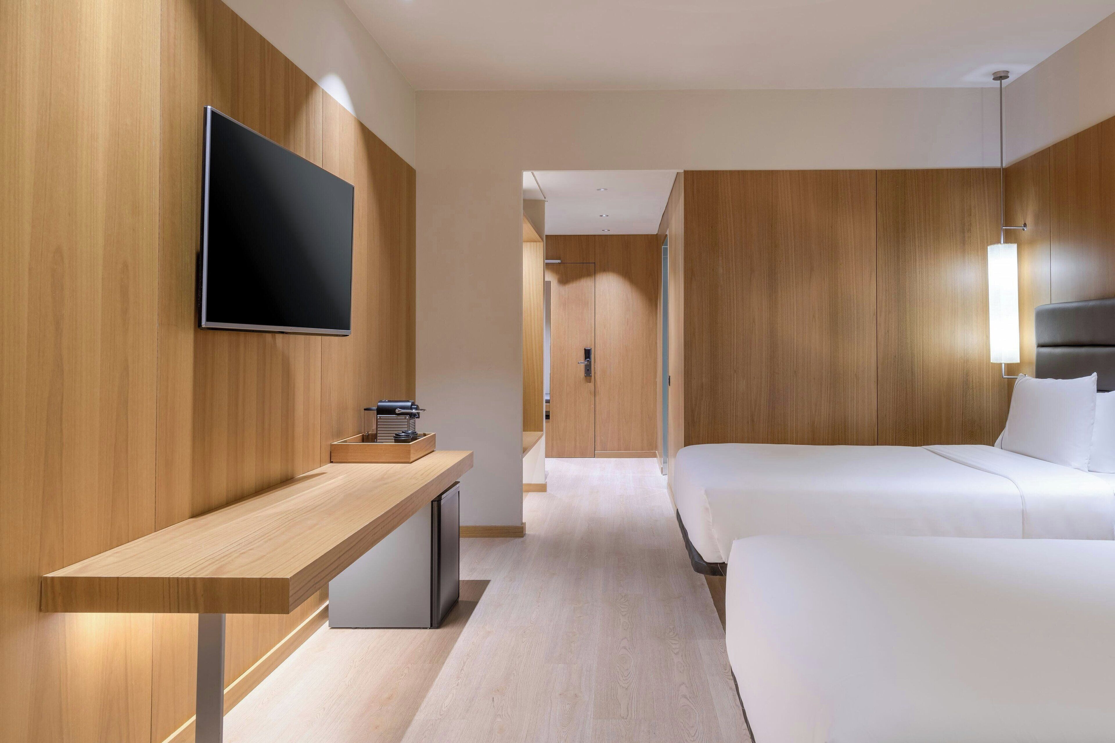 Una habitación de hotel moderna con una pared de madera, un televisor de pantalla plana sobre un escritorio de madera, una cama con ropa de cama blanca y una mesita de noche.