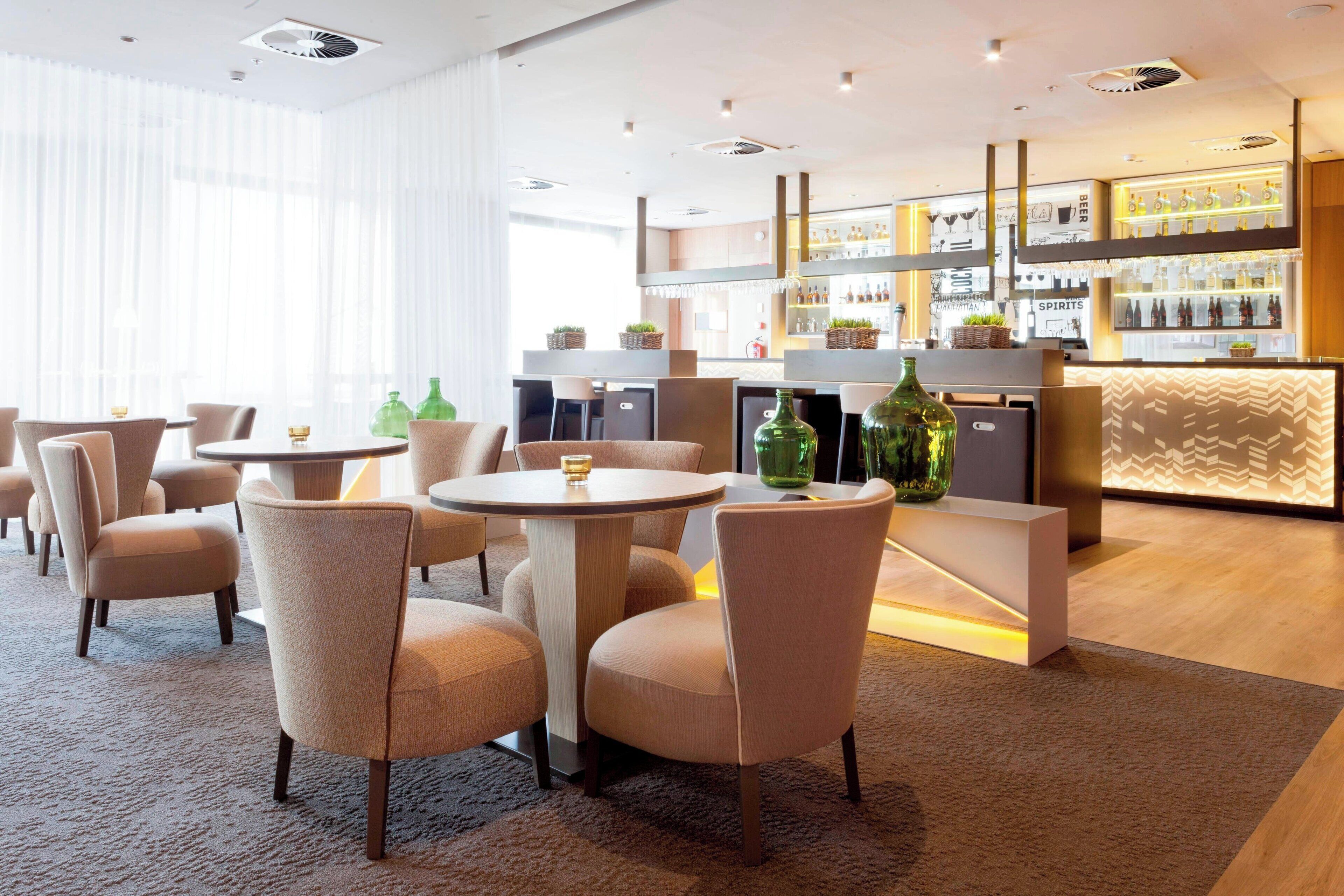 A modern bar area with round tables and chairs, a bar counter with shelves of bottles, and a patterned wall in the background.