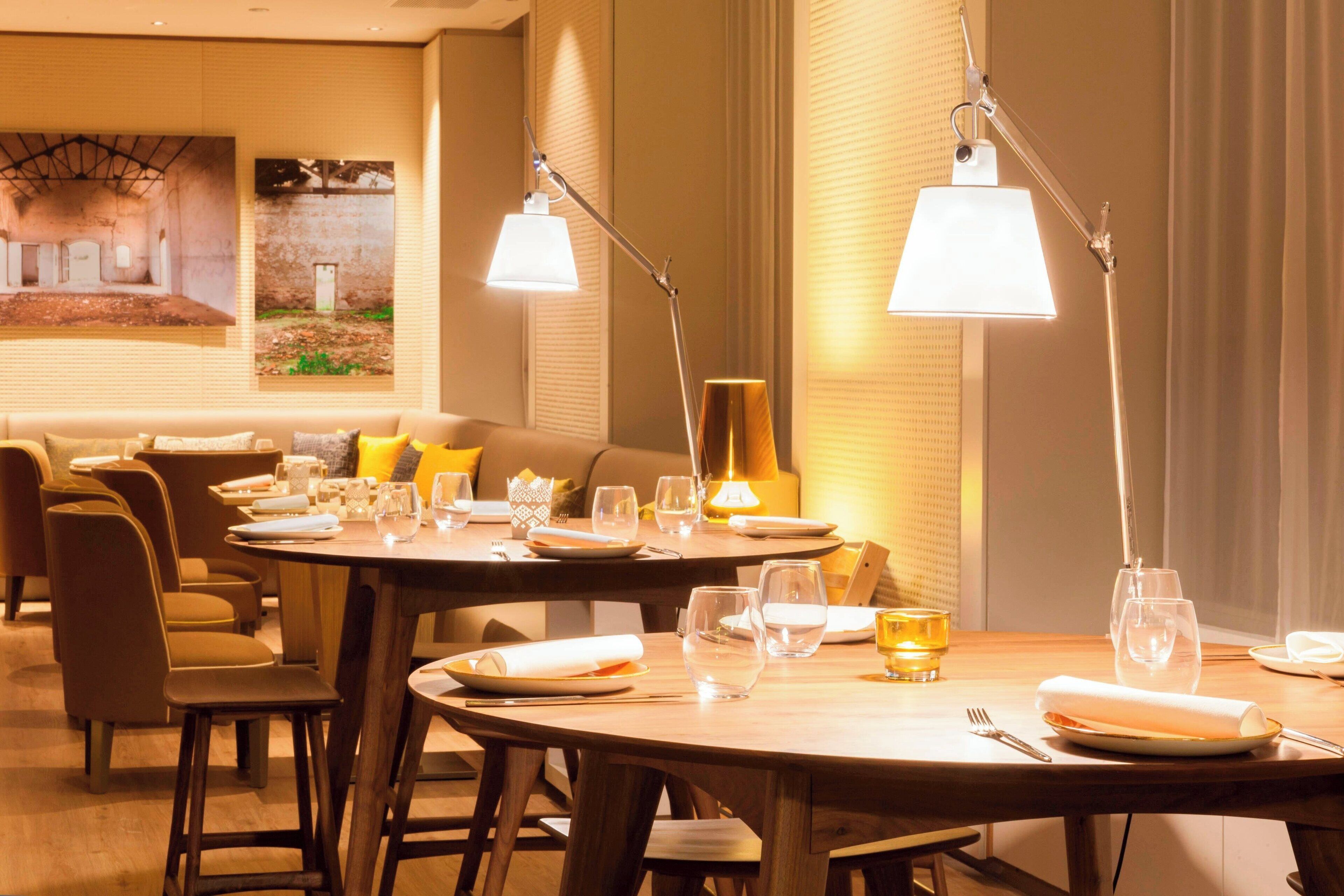 A dining area with tables set for four, each with plates, glasses, and cutlery. There are two pendant lights above the tables, and a sofa with cushions in the background.