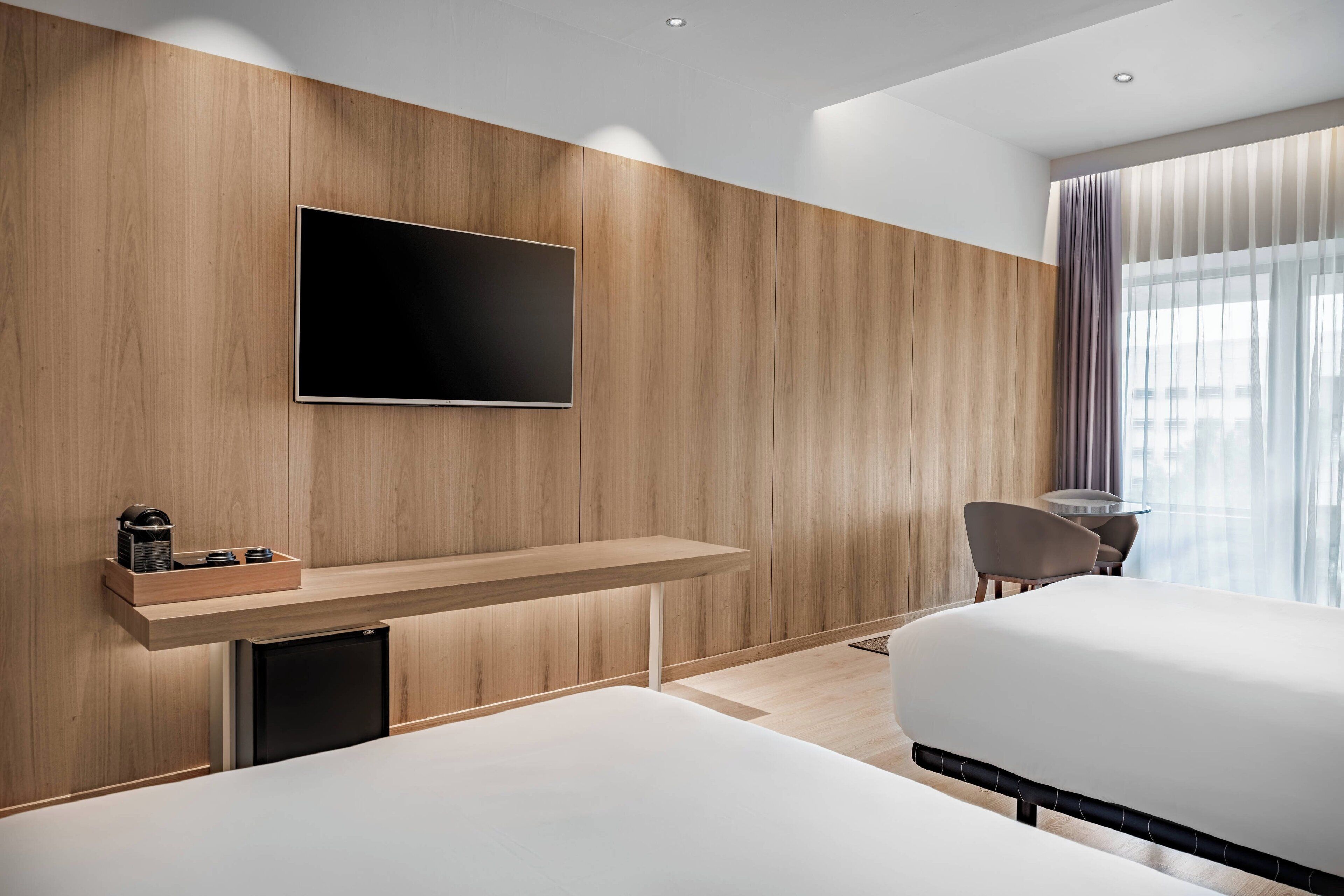 A modern hotel room with a wooden panel wall, a flat-screen TV, a small desk with a coffee maker, and a bed.