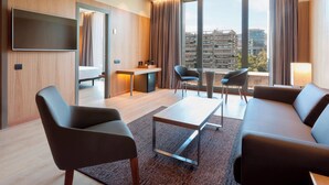 A modern hotel room with a large window, a flat-screen TV, a sofa, a coffee table, and a desk with a chair. - AC Hotel Diagonal L'Illa by Marriott (Barcelona)