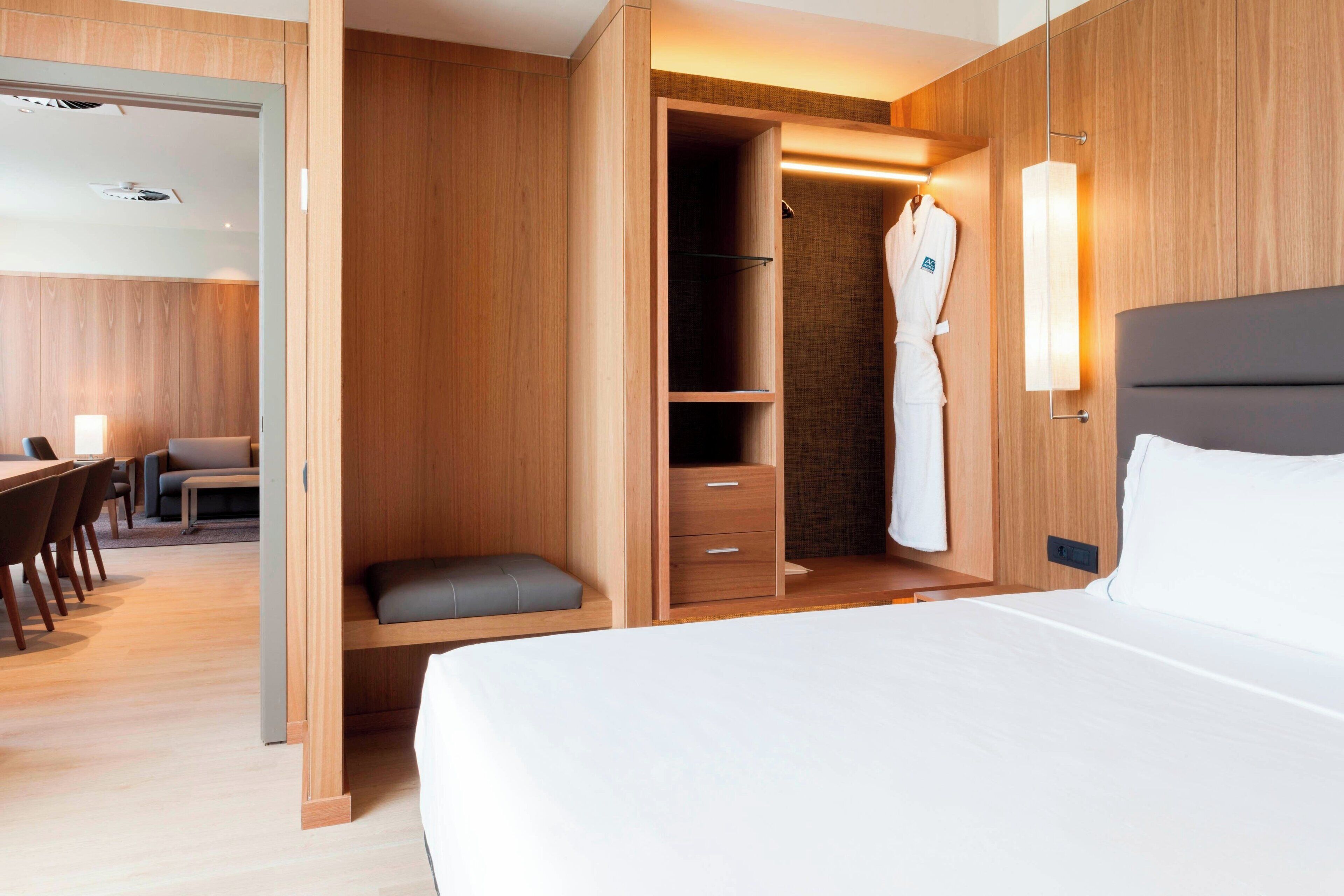 A modern hotel room with a large bed, a wooden wardrobe, a seating area with a table and chairs, and a robe hanging on the wall.