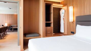A modern hotel room with a large bed, a wooden wardrobe, a seating area with a table and chairs, and a robe hanging on the wall.