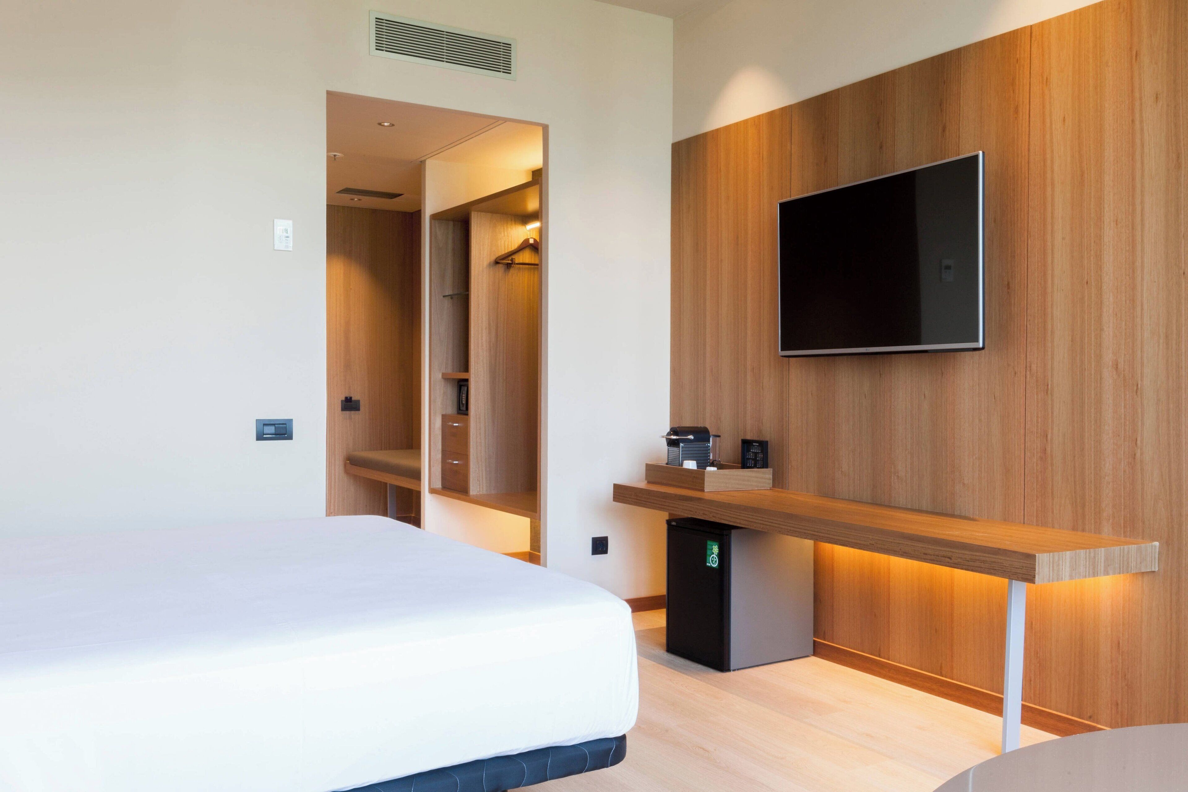 Een moderne hotelkamer met een bed, een aan de muur bevestigde televisie, een klein bureau met een koffieapparaat en een inloopkast.