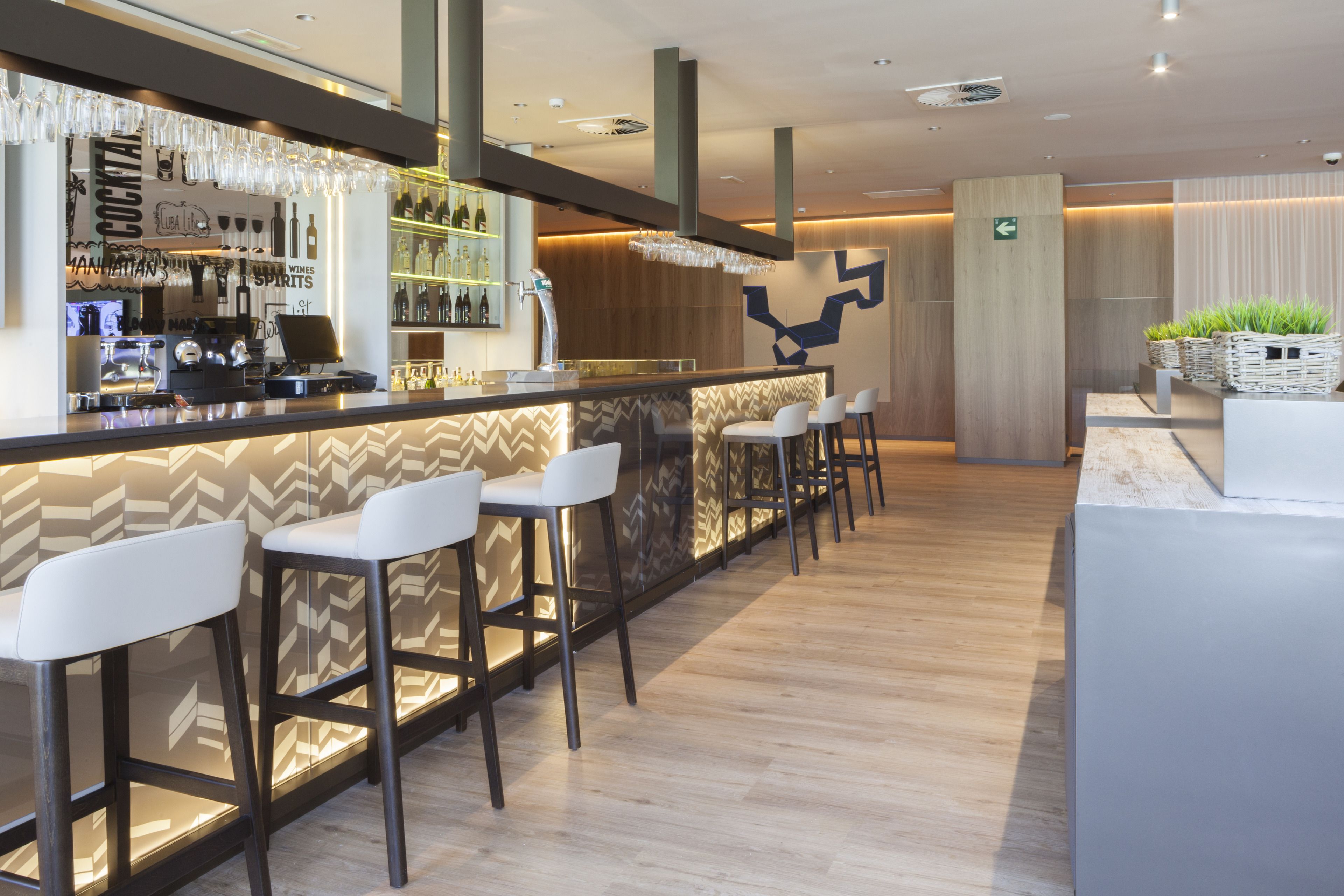 A modern bar area with a bar counter, white bar stools, and a selection of bottles and glasses.