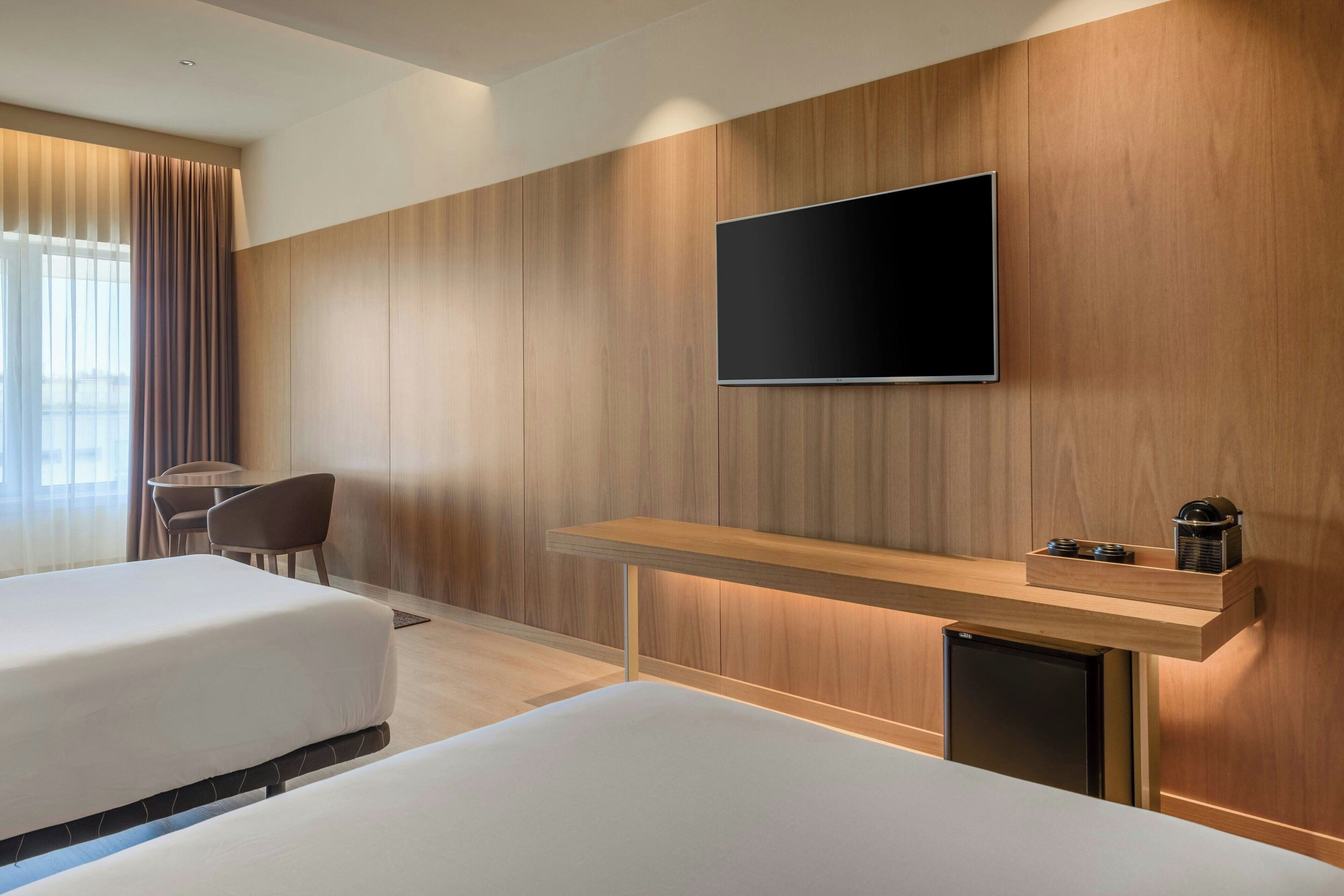 Habitación de hotel con dos camas, un televisor montado en la pared, una mesita con cafetera y una silla.