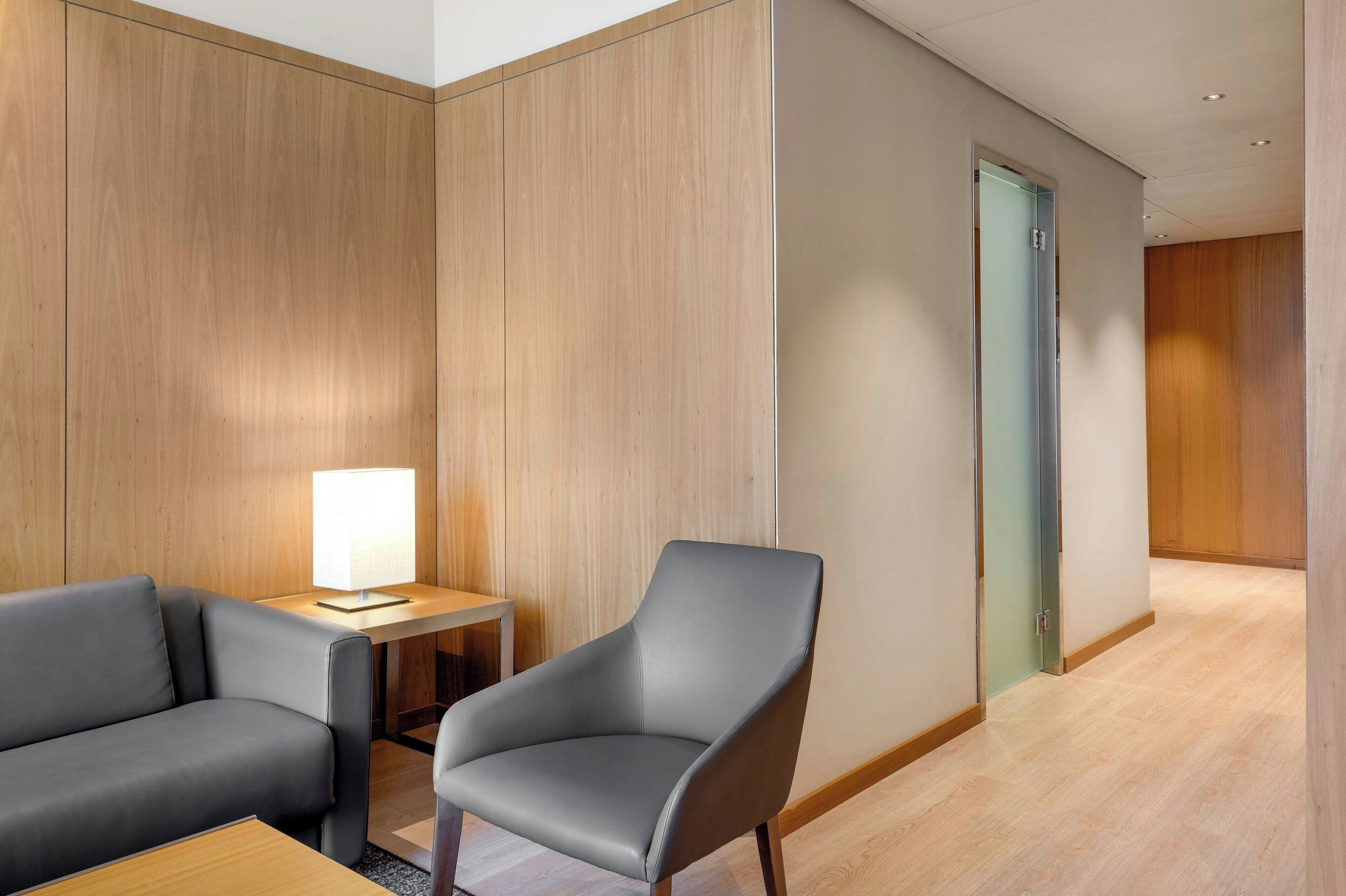 A hotel room with a wooden panel wall, a grey armchair, a small table with a lamp, and a door leading to another room.