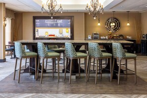 A bar area with a long counter, six patterned chairs, a television screen, and a chandelier.