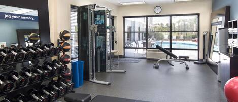 Une salle de sport équipée de matériel de musculation, d’un banc et offrant une vue sur une piscine grâce à de grandes fenêtres.