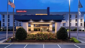 Eingangsbereich eines Hampton Inn Hotels mit überdachtem Außenbereich, amerikanischen Flaggen und einem Parkplatz.