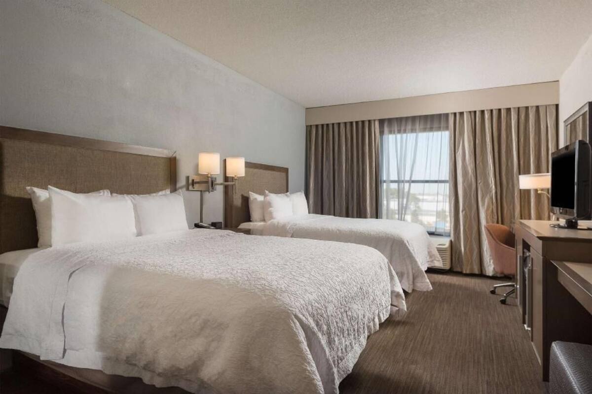 A hotel room with two beds, a desk with a television, and a large window with curtains.